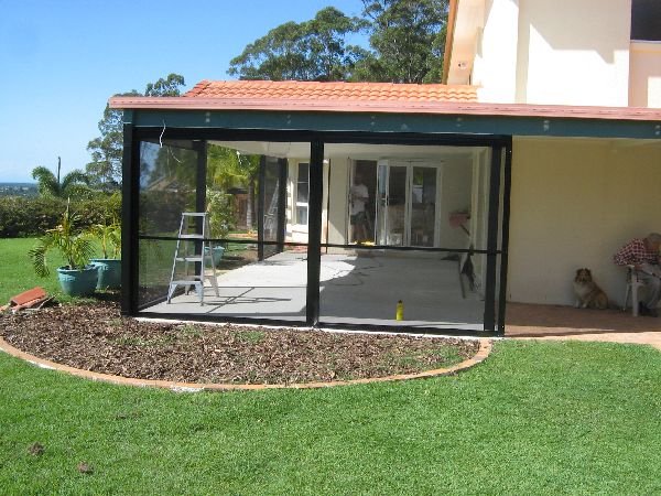 Patio with dark frame and glass walls attached to a house with a red-tiled roof. The patio floor is unfinished, and there's a ladder inside. A small garden with mulch is in front, and a person sits near a dog to the right.